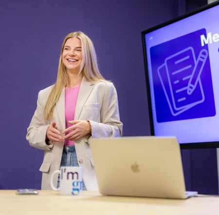 Anne-Wil geeft een training over Meta Ads. Ze staat lachend voor een beeldscherm met een slide over Meta Ads opzetten.