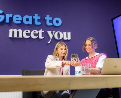 Eileen de Jong en Anne-Wil prange kijken op een telefoon voor een paarse muur waarop 'Great to meet you' staat.